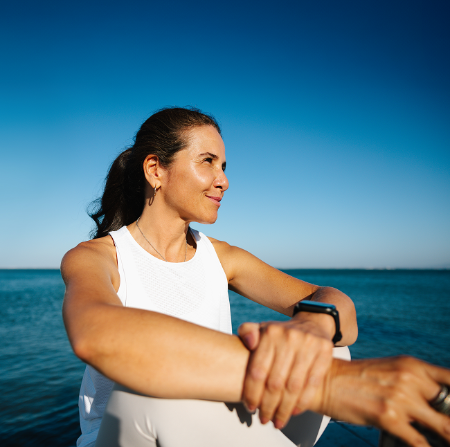 Woman stretching sitting by the ocean on a clear day, wearing a white athletic top and fitness watch.